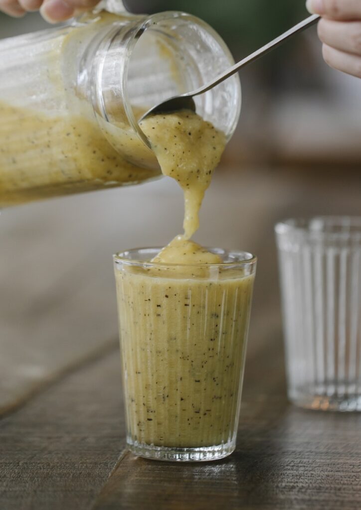 Crop unrecognizable person pouring tasty banana smoothie into glass placed on wooden table